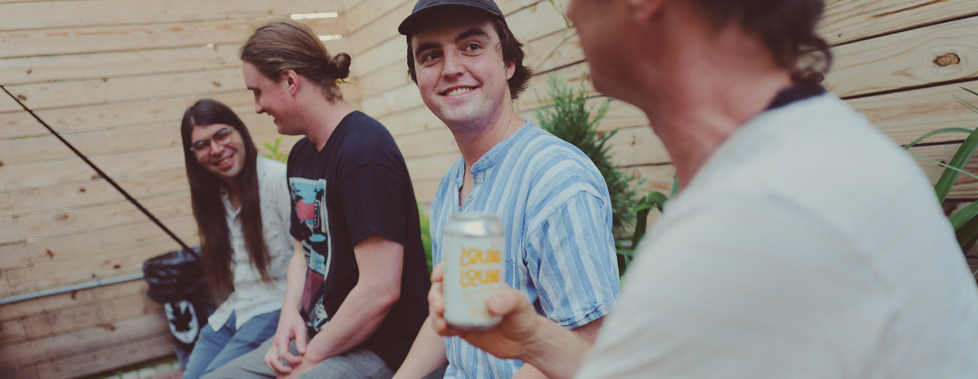 Four friends sitting together, one holding a can, in an outdoor setting.